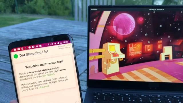 Still from the video: my phone, running the Dat Shopping List demo and my computer showing the desktop wallpaper. The video is shot in the garden with a  wooden table and chairs and grass in the background.
