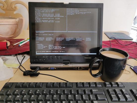 OpenBSD laptop in ttyC1 running Tmux. Top left pane is running mpv streaming aNONradio.net.  Top right pane is showing output from mixerctl. Bottom pane is SSHed to SDF and showing output from anonradio chatroom in commode. Second cup of coffee with half-&-half to the right of the screen. Keyboard peeking from bottom of image.