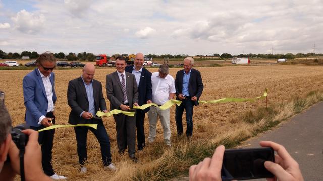 6 Prominente durchschneiden symbolisch ein Band, dahinter der erste Bagger auf einem Tieflader der dann mit den Arbeiten anfängt. Viel freies Feld.