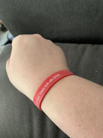 Rechter Unterarm mit Faust vor einer Couch. Und einem roten Armband beschriftet mit „Lieber Tanz ich als D20“