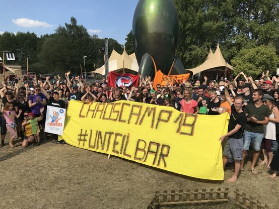 Menschen mit Transpi "Chaoscamp 19 #unteilbar" vor CCC-Rakete.