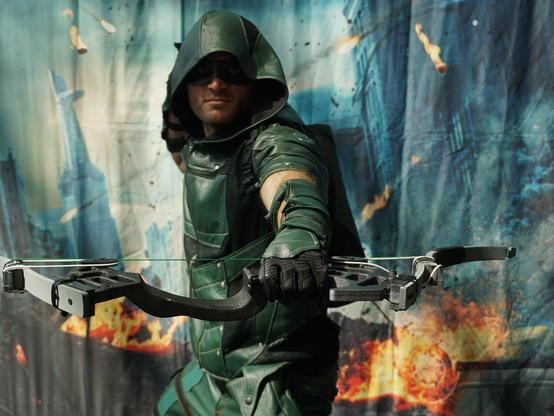 Cosplayer dressed as Arrow-style Green Arrow against a backdrop of a disaster-struck city.