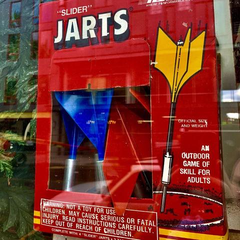 A box of “JARTS” sits in a window display. This is a rebranding of the generic Lawn Darts game, complete with a large warning reading “Warning: Not a toy for use by children. May cause seriou
