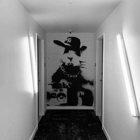 A long, white hallway. Fluorescent bar bulbs adorn the walls at angles. At the end of the hallway, a full-wall stencil of a rabbit with an ‘80s boom box, gold chain, and sideways ball cap