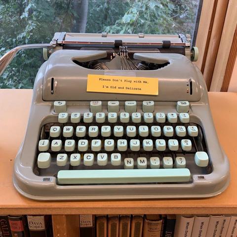 A typewriter sits on a bookshelf. Many of the keys are stuck down and it looks non-operational and decorative. Above the keyboard is a small yellow sign with typed text that reads, “Please do