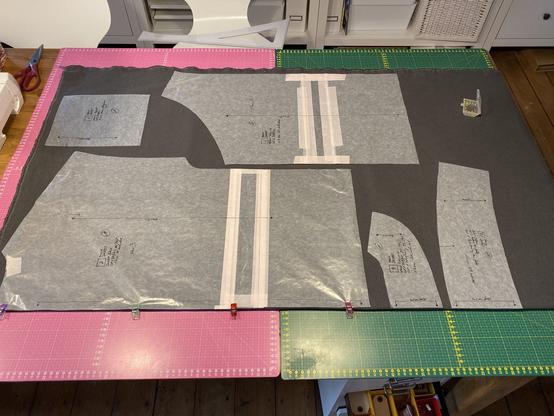 Ein Tisch mit Schneidematten, auf dem sich eine gefaltete Bahn mit dunkelgrauem Cordstoff liegt. Darauf verteilt sind Schnittmusterteile aus Pergamentpapier verteilt.