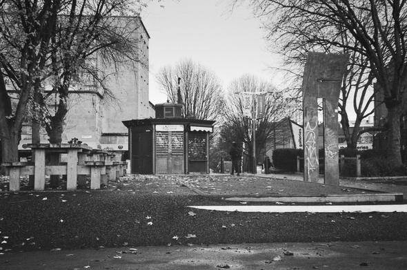 A monochrome photo of Edna's Kitchen - an octagonal turret type cafe on the edge of Castle Park, Bristol, UK. With associated tables and seating on the left. People can also be seen, going on their way.