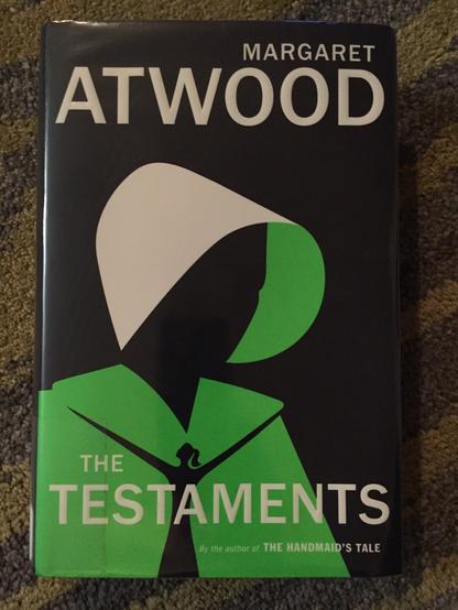 picture of a hardcover copy of margaret atwood's novel THE TESTAMENTS, with a green-clad, bonnet-hooded female figure on the cover