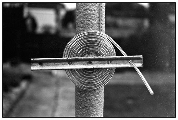 Monochrome photograph of a plastic coated steel cable, coiled around a horizontal stay bar fixed to a scaffolding pole that has been painted with Silver Hammerite.