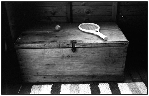 A monochrome photograph of an old foot locker or wooden box in a garden shed. A striped rug can be seen on the floor. There a child play racket and ball on the lid of the locker.