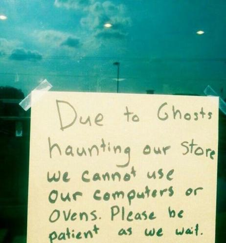 A handwritten note taped to a storefront window that reads "Due to Ghosts haunting our store, we cannot use our computers of ovens. Please be patient as we wait." Gloomy clouds are reflected  by the glass behind.