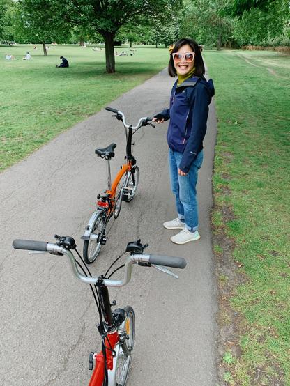 A beautiful girl with pink shade walking on a park path with her orange bicycle. Another bike, pushed by the photographer, is in the foreground.