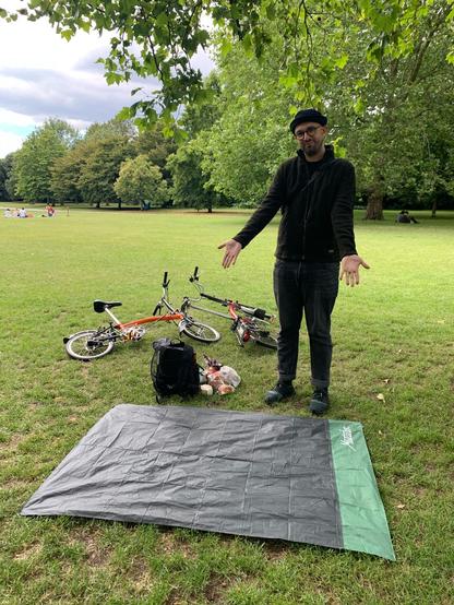 A high tech, highly foldable, highly packable picnic blanket has been deployed on the ground in a park. The author can be seen proudly gesturing towards it. Two bicycles are on the ground in the background.