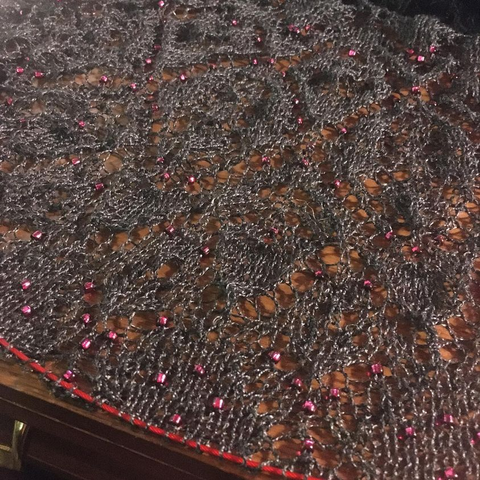 A knit lace shawl in process. The yarn is a silvery grey, and scattered with dark red beads.