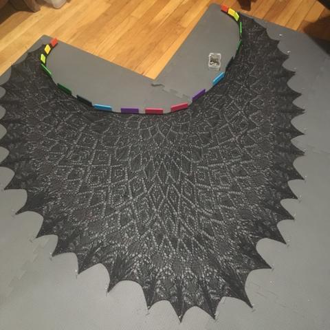 Blocked knit lace shawl, made from black yarn and scattered with red beads. It is pinned in place.