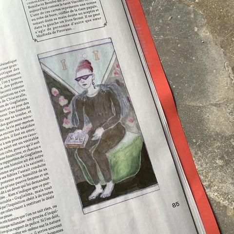 La Papesse vêtue d'un justaucorps noir; elle a les pieds nus et les épaules recouvertes d'une pèlerine bleue retenue par 2 fibules. Elle est assise sur un pouf vert, elle porte des lunettes de soleil aux verres triangulaires, et un triple chignon un peu punk (rasé sur les côtés) surmonté d'une sorte d'oeuf blanc. Elle a une clé qui pend de son oreille.