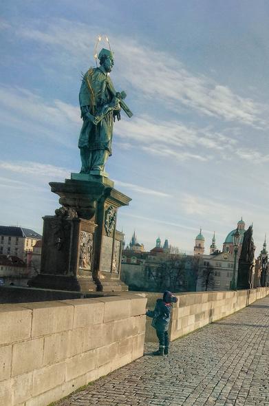 Enfant levant la tête vers une statue baroque représentant Saint Jean Nepomucène en habits de prêtre, tenant un crucifix. Temps froid et ensoleillé, clochers baroques en arrière plan, la photo est prise sur un pont de pierre du XVè siècle.