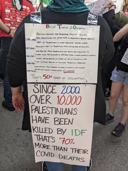 A woman wearing hijab has a poster with a handwritten timeline of the occupation on her back. Below the timeline is a sign that reads "Since 2000, over 10,000 Palestinians have been killed by IDF. That's 70% more than their COVID deaths"