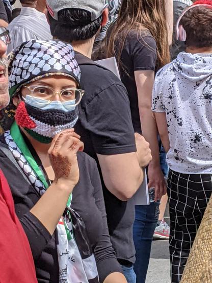 A protestor wearing a crocheted mask in the pattern of the Palestinian flag over a surgical mask looks into the camera. The demonstrator has clear glasses and a silk headscarf with a keffiyeh design.