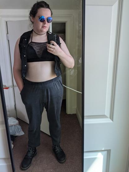 Mirror selfie of a white woman with her dark hair in a bun wearing a black denim vest over a black halter top and black sweatpants worn at the natural waist, accessorized with black boots, a tattoo choker, and circle-rim reflective glasses. I stand casually, with one hand in my pocket.