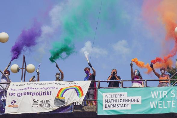 Auf dem Bild ist ein Transpi an einer Brücke zu sehen mit der Aufschrift: "Fast alle Superreichen sind hetero-maker. Vermögensabgabe ist Queerpolitik." Darunter sind die Logos von Attacikka und Wer hat der gibt zu sehen, daneben ein Topf am Ende eines Regenbogens, der wie ein Pfeil vom Gold weg geformt ist. Auf der Brücke stehen Menschen die bunten Rauch halten.