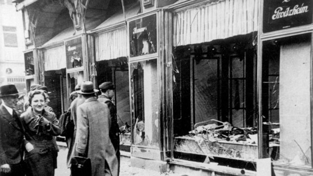 A black & white photograph shows the aftermath of Kristallnacht, 9--10 November 1938, when Nazi SA thugs smashed and looted Jewish shops, buildings, and synagogues throughout Germany.

Shop windows stand smashed, glass litters the street, passersby look on, a man and woman facing the camera both smile.