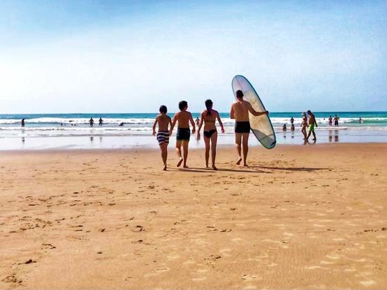 Landscape picture of a large and not too populated beach on a bright and sunny day. The focus of the photo is an adult carrying a surfing board with three kids at his side walking purposefully toward the ocean.
