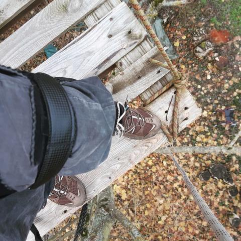 Das Bein einer Person ist, zu sehen,  sie trägt eine Bandage (Schiene aus achwarzen Plastik). Sie trägt weiter Wanderschihe. Die Person steht auf einer Holzplatform in luftiger Höhe in einem Baum, weit unten ist laub zu sehen