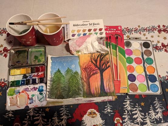 Painting and painting equipment spread out across a table covered with a cloth with a Nordic winter / Yuletide scene of Julenissen eating porridge with a wooden spoon from a bowl as a kid holding presents looks at him with joy.
The painting is a diptych of a cold starry winter night scene with snow falling down on trees, and a warmish fall scene of bare trees and tall grass bathing in a golden orange sunset glow.
The painting is surrounded by the water colour painting equipment used to create it: a small metal case with half-pan water colour paints, Panduro Watercolour Basic Set; brushes I borrowed from the kid's Christmas present; a couple of sheets of toilet paper to dry the brushes, coloured by the paints used; a set of more opaque water colour cakes; and two paper cups with the dirty water from cleaning the brushes (one for 'cold' and one for 'warm' colours).