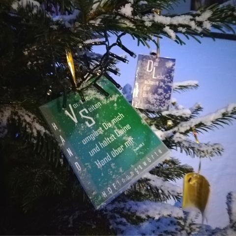 Schneebedeckte Tannenzweige. Vorn hängt an einem Zweig ein grüner Zettel mit der Aufschrift "Von allen Seiten umgibst Du mich und hältst Deine Hand über mir. Psalm 139,5