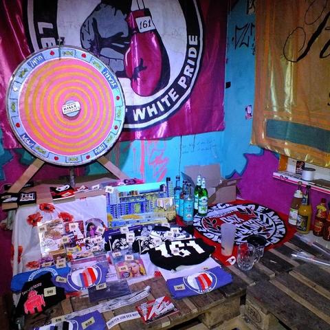 Das Bild zeigt ein Glücksradstand mit diversen Preisen aus T-Shirts, Sticker, CDs, DVDs, Schallplatten, Aufnäher, Bücher und Trost-Schnaps/Saft.
Im Hintergrund hängt eine rosa Fahne mit "Let's fight white pride-logo" mit Boxhandschuhen.