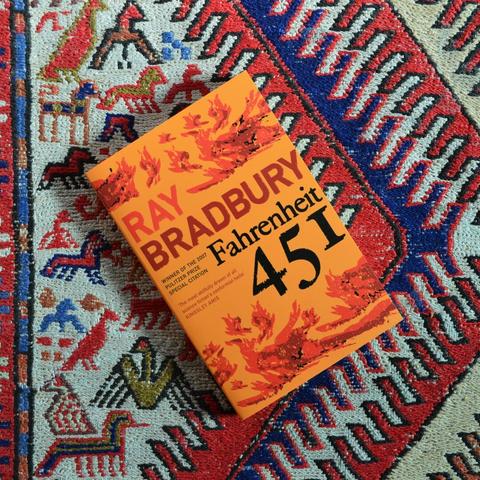 A copy of Ray Bradbury's Fahrenheit 451 lying on a Turkish rug. The book has an orange cover with red flames on it. The title is black and the author's name is red.