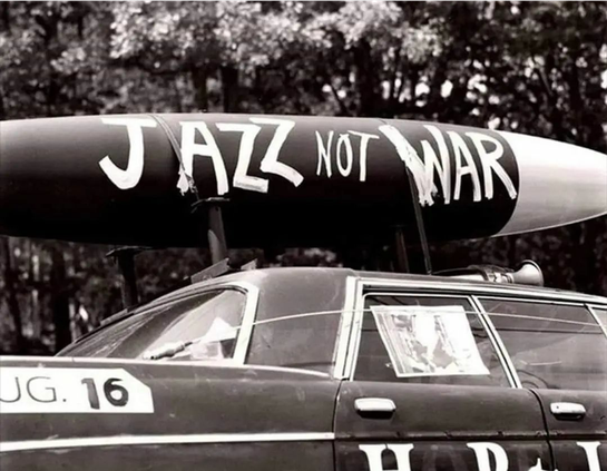 Photo en noir et blanc, probablement des années 60.
Une voiture porte une fusée avec le slogan peint : jazz not war