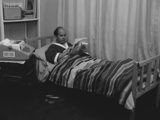 Old 60s BBC doc. Middle-aged man, in housecoat, lying in bed, reading  papesr from a teletype writer installed next to his bed.