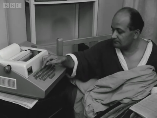 Old 60s BBC doc. Middle-aged man, in housecoat, lying in bed, inputting commands into a teletype writer installed next to his bed.