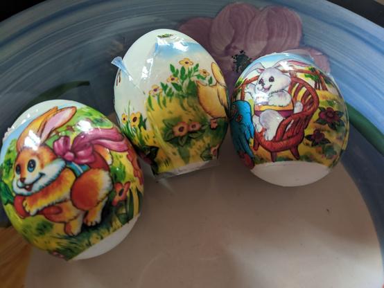 Three eggs with 3 different designs applied.
First one shows an easter bunny holding a carrot, with a bow around its waist.
Second shows a chick butt, grass and flowers, and that the wrap is slightly torn at the top of its seam; it's also not nicely centered around the egg because of it.
Third is another bunny, sitting on a chair at a table.