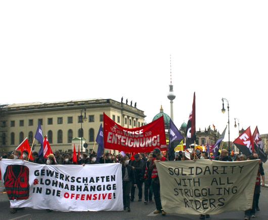 1. Mai-Demo in Berlin Mitte, mit dem Fernsehturm in Hintergrund. Auf dem Foto sind viele Menschen mit FAU-Flaggen und Demo-Transparenten zu sehen, z.B.: Gewerkschaften & Lohnabhängige in die Offensive! – Von der Krise zur Enteignung! – Solidarity with all struggling riders!