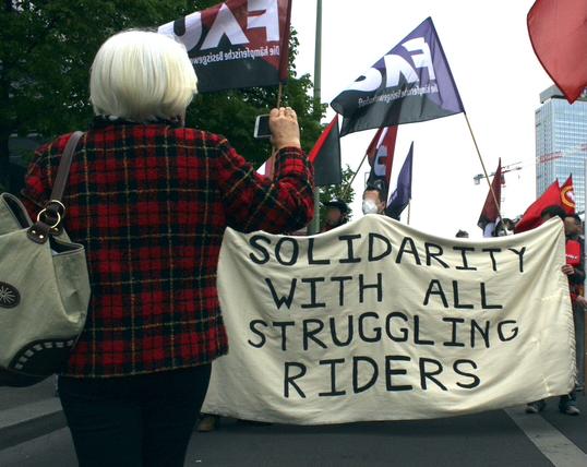 Foto von der 1.Mai-Demo mit dem Transparent: "Solidarity with all struggling riders". Im Hintergrund sind rote und lila FAU-Fahnen. Vorne im Bild sieht man eine ältere Dame, die ihr Smartphone in Richtung Demo hält.