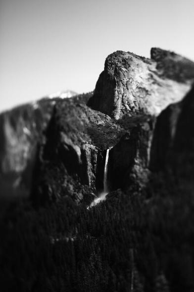 a black and white landscape photo of bridalveil falls in yosemite valley. taken from a great height, overlooking the valley. the waterfall flows in the distance with pine trees covering the valley floor and mountain tops.