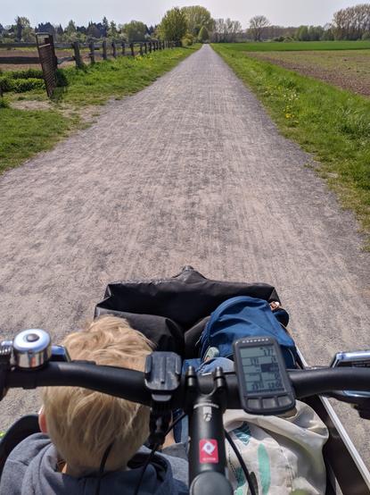 Das Bild zeigt aus Sicht des Fahrers den vorderen Teil eines Bullitt Lastenrads unterwegs auf einem Feldweg. In der Kiste sitzt ein Schulkind, neben ihm sein Schulranzen, Turnbeutel und eine Einkaufstasche. Die Sonne scheint.