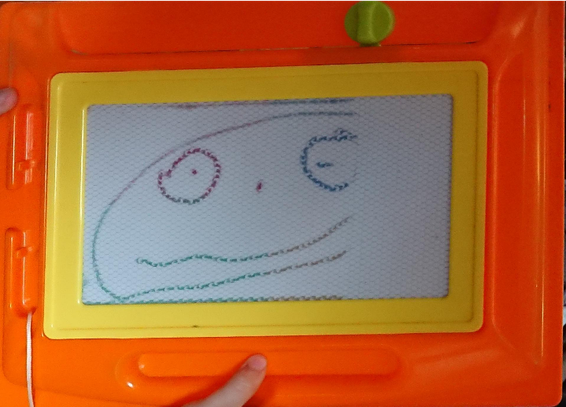 a simple face with two round eyes with dots for pupils, a dot as a nose, and a squiggly line as its mouth in a somewhat smiling pose. The right-side of the face is slightly missing as the magnetic sketch-board's erase slider slid to the left as kiddo was holding it up.
