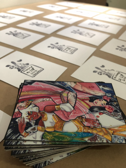 A lot of cards, with the back upside. Stamps and Numbers are on it.
In the front are two girls, one in a kind of kimono and dark hair. Wearing flowers. The other one wearing gold and white dress. Her white hair and Cat ears frame her dark skin.
Both are upside down to each other, like a gaming card