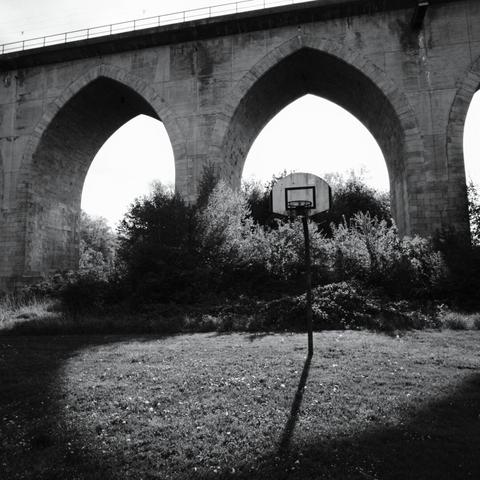 Foto einer hohen Eisenbahnbrücke mit gemauerten Pfeilern. Im Vordergrund ist eine Wiese mit Basketballkorb. Die Brücke wirft Schatten in Richtung Fotograf,   mit der Ausnahme eines halbrunden Bereichs um den Basketballkorb, durch Licht dass durch einen der Brückenbögen fällt.
