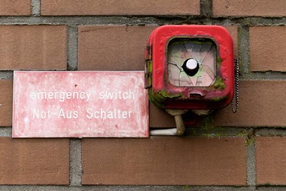 Ein verrosteter, mit Moos bewachsener Druckknopfmelder. Die Scheiben ist gebrochen. Auf einer Ziegelwand. Ein Schild daneben :"emergency switch, not-aus Schalter"