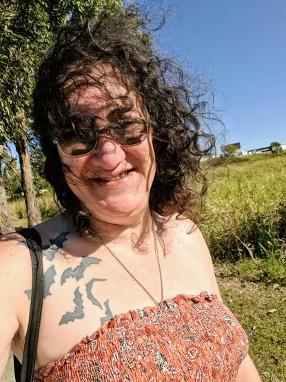 A selfie showing a smiling woman wearing tinted glasses. Her shoulder length curly brown hair is blowing across her face. A field of green grass is in the background