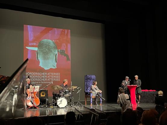 Foto der Bühne, die Preisträgerin sitzt auf dem Thron. Die beiden Laudator:innen „bei der Arbeit“.