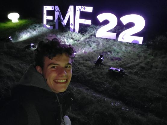 A picture of me standing outside a lit up EMF22 sign on the campsite.