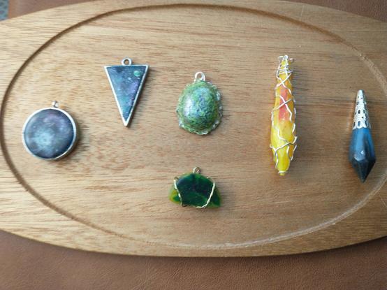 A wooden platter filled with pendants, including a crystal shaped yellow pendant with a red orange center highlight and silver wire wrapped around it; a circular galaxy pendant; an upside down triangle pendant with planets and stars; a green, black, and copper entanglement pendant with wire wrapping; a crescent shaped pendant wrapped with gold wire and colored with yellow and green blooming in the center; and black and blue pendant with a silver cap.