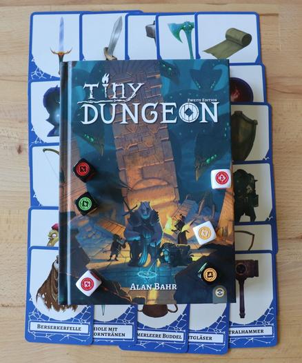 Tiny Dungeon Regelwerk auf einem Bett aus Ausrüstungskarten. Darauf verteilt sechs Würfel.