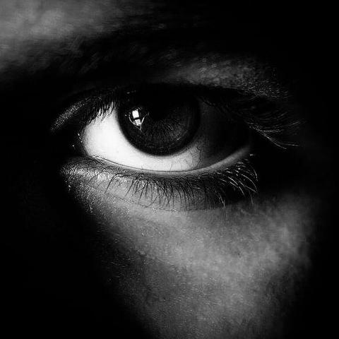The black and white photo shows a close-up of a human eye, the eyelashes. The rest of the face is veiled by shadows.